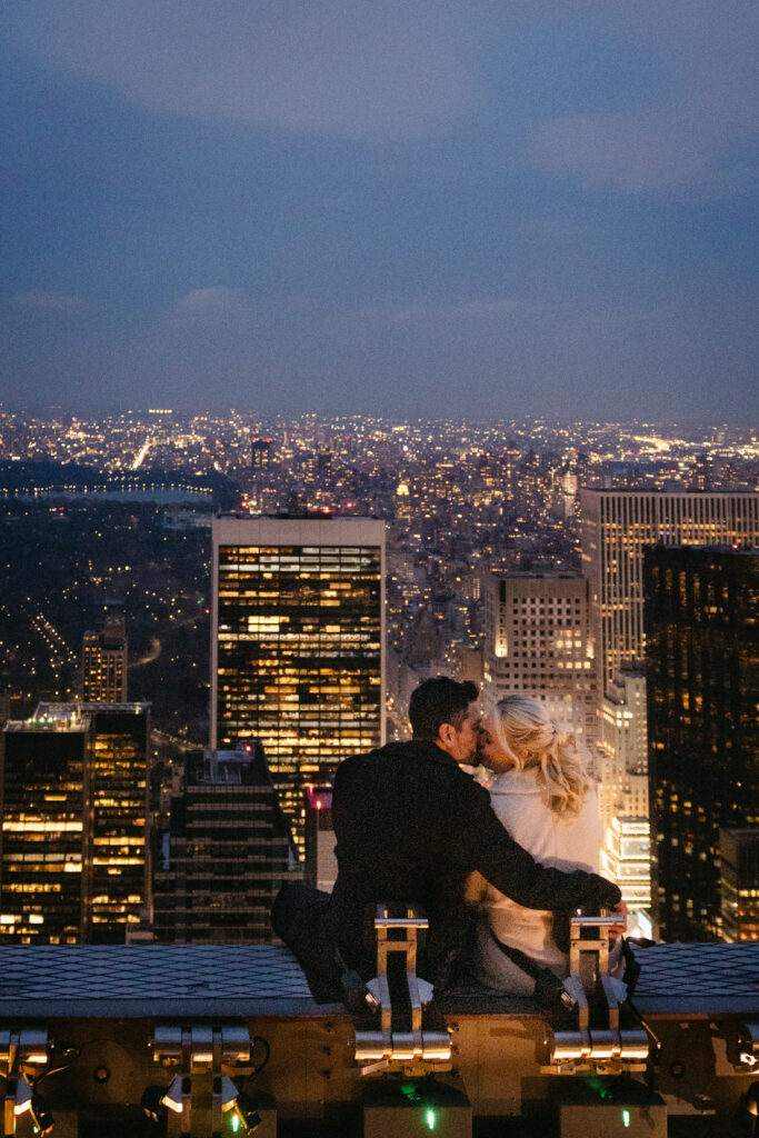nyc elopement couple sitting on a beam at the top of the rock with nyc skyline in the background