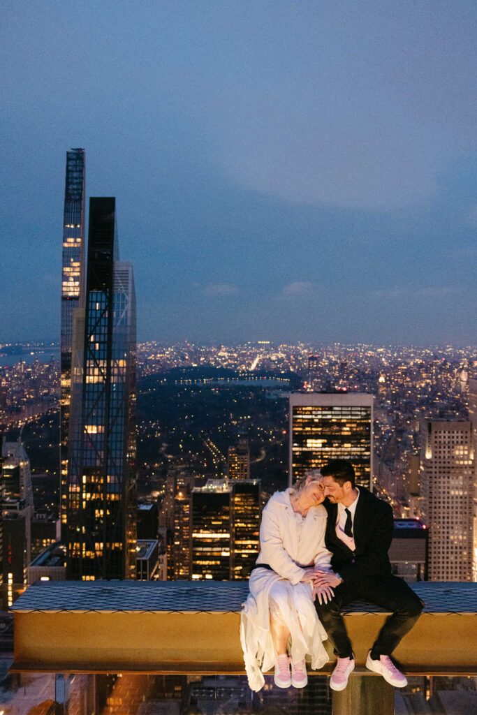 nyc elopement couple sitting on a beam at the top of the rock with nyc skyline in the background