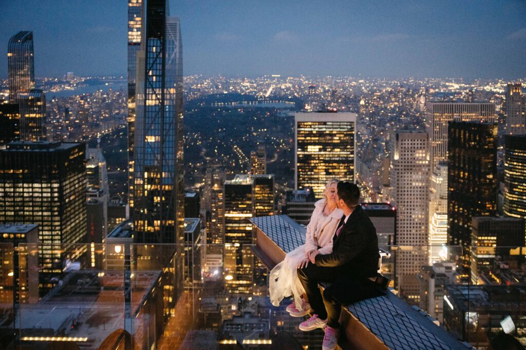 nyc elopement couple sitting on a beam at the top of the rock with nyc skyline in the background